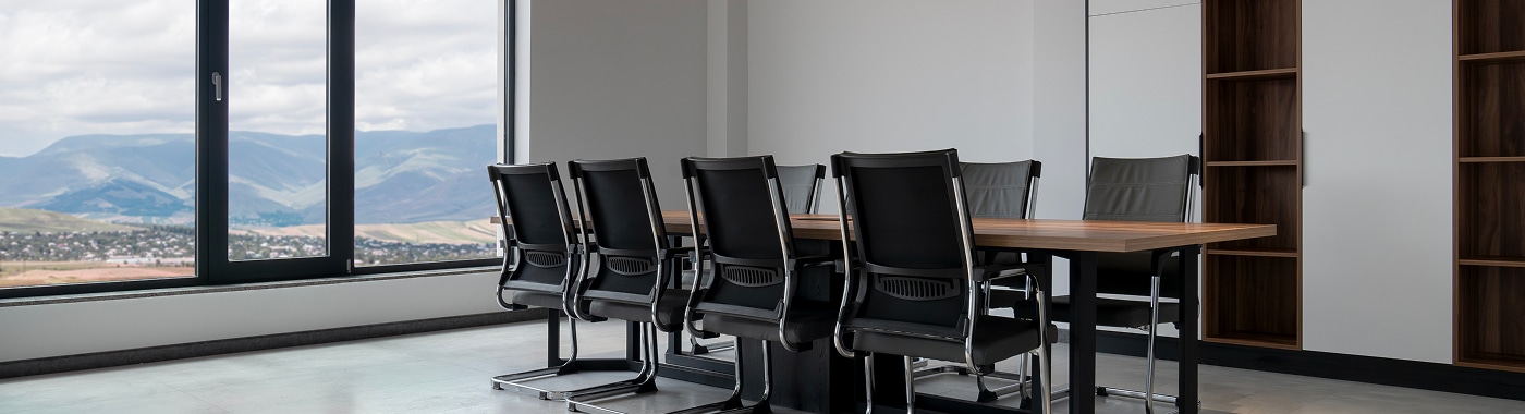 A modern, empty conference room featured on the About Us Page of Massaro Law Firm, with a wooden table, eight black chairs, built-in shelves, and large windows revealing mountains and a cityscape outside.